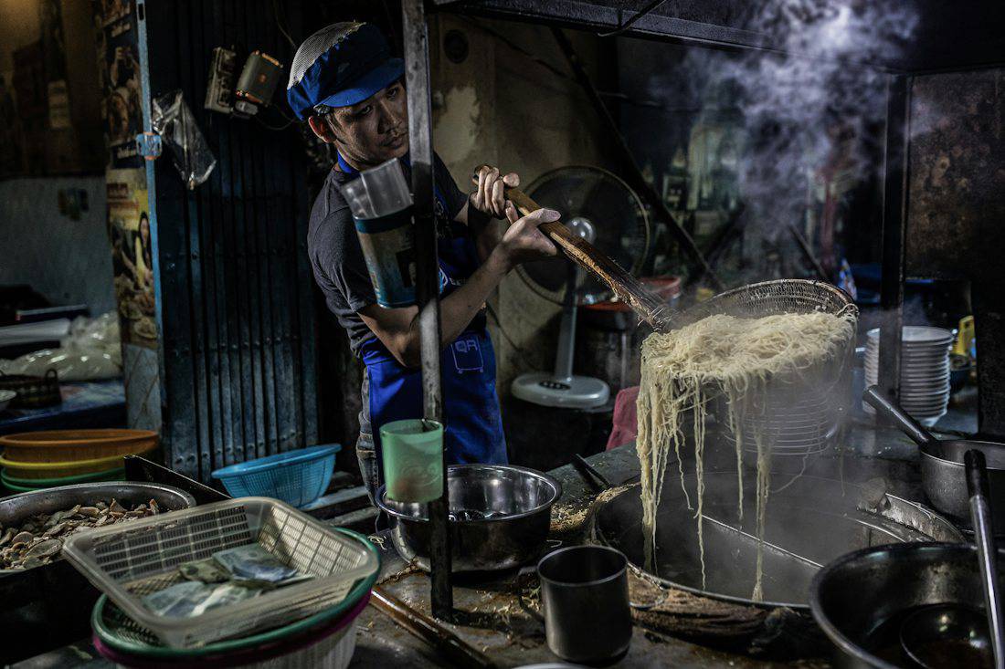 Cook preparing noodles in a steaming wok at a Bangkok street kitchen