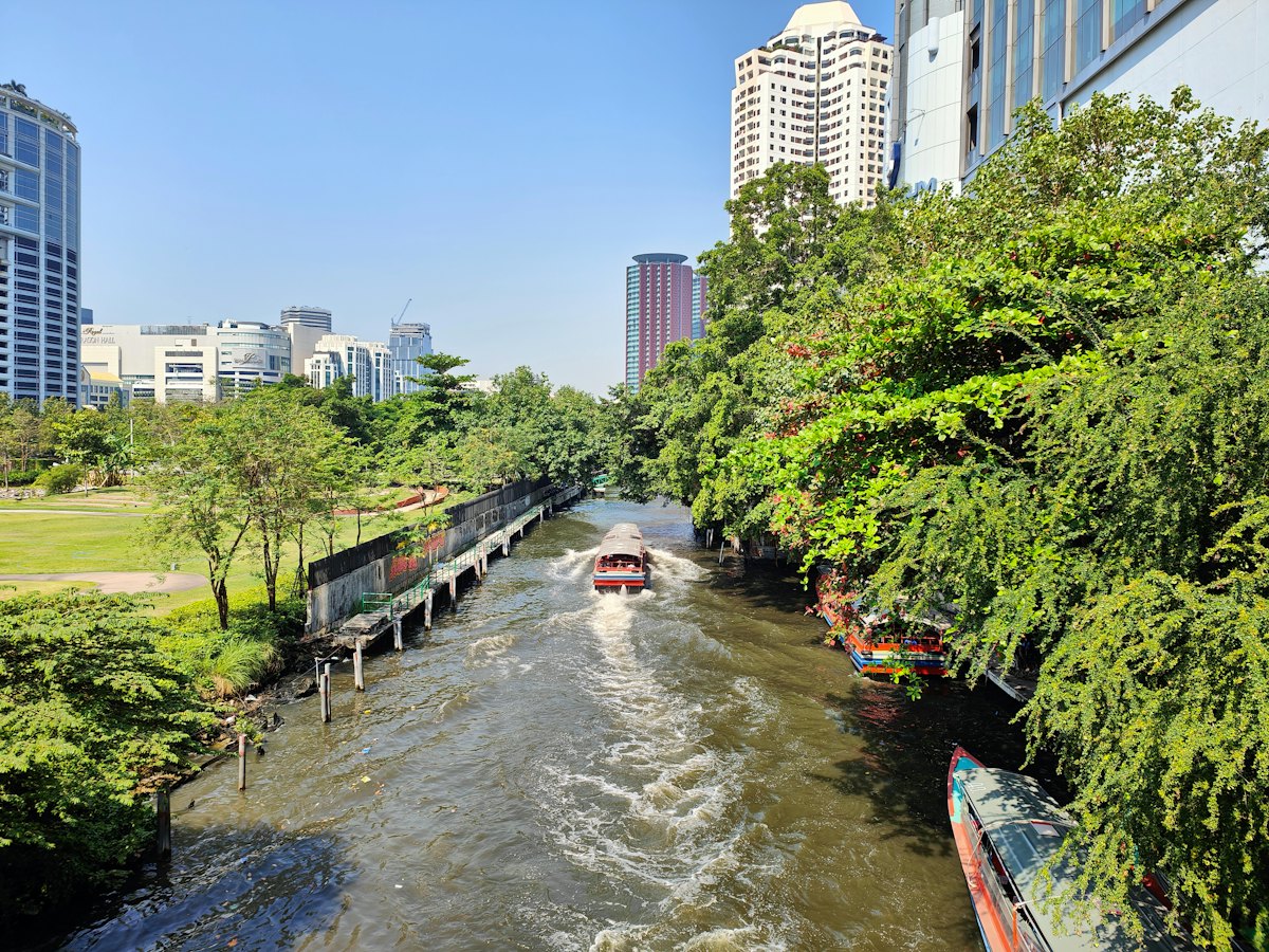 A canal passenger boat speeding along a tree-lined khlong between Bangkok towers
