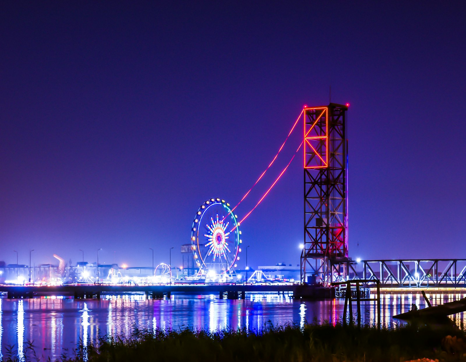 A lit ferris wheel and tower ride on a riverfront at night