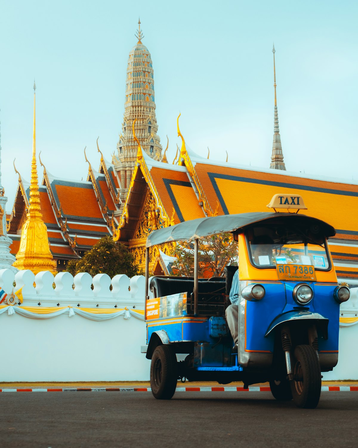 A blue and yellow tuk-tuk parked on the road beside the outer wall of the Grand Palace in Bangkok