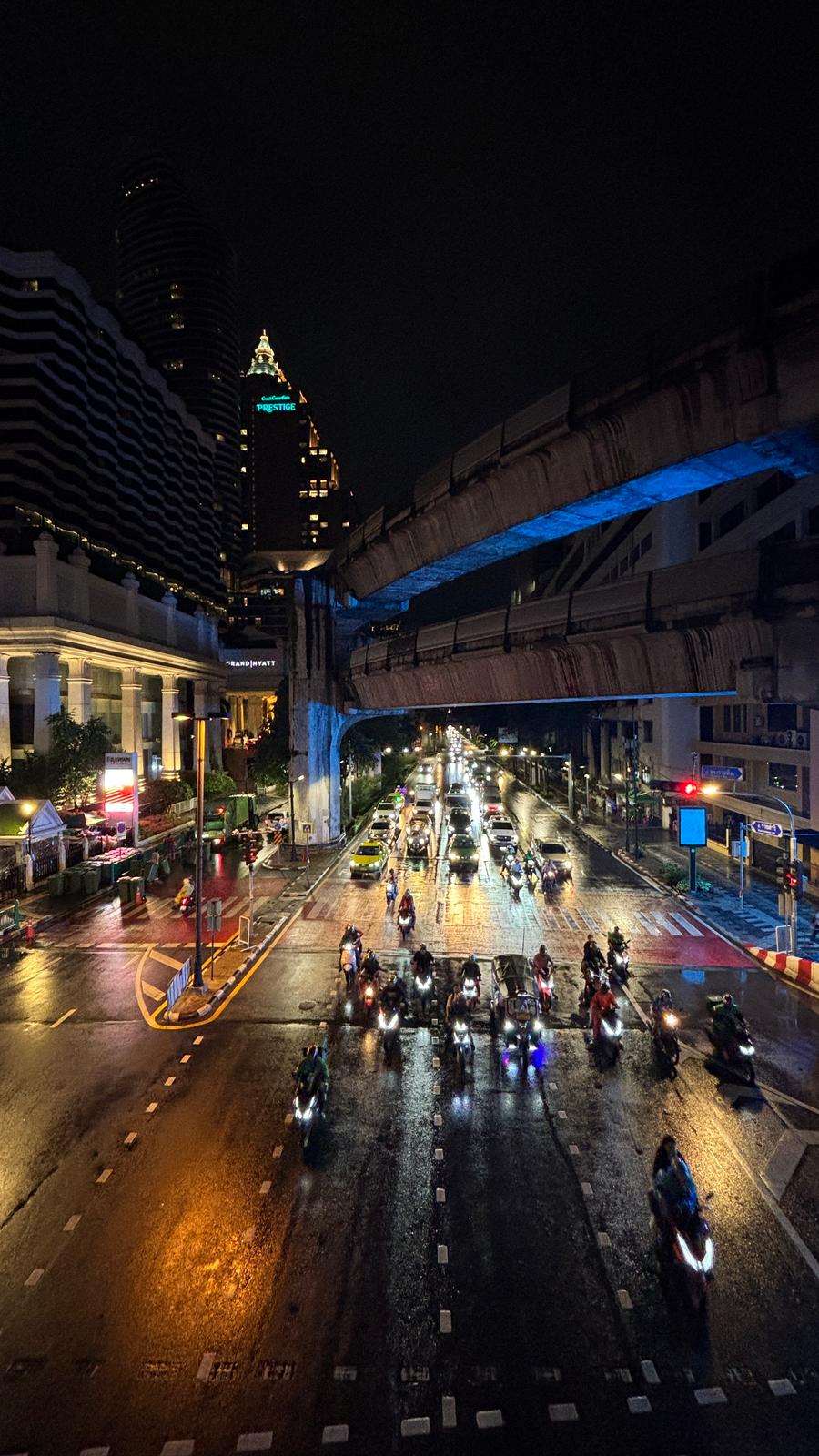 Cars and a wave of motorbikes on a wet road at night under the BTS viaduct at Ratchaprasong, Bangkok